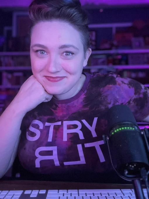 Emily Friedman in a low lit room, hair up, with an ice-dyed black and magenta shirt that read STRYTLR in large white capital letters.