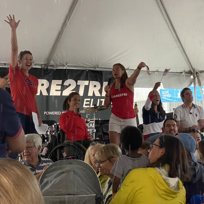 Paralympic athletes on stage under white tent with arms in the air and Dare2Tri Elite banner in background