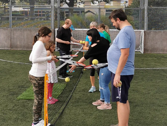 A group of men, women, boys and girls hold tennis rackets with soft yellow balls on over a pop up net.