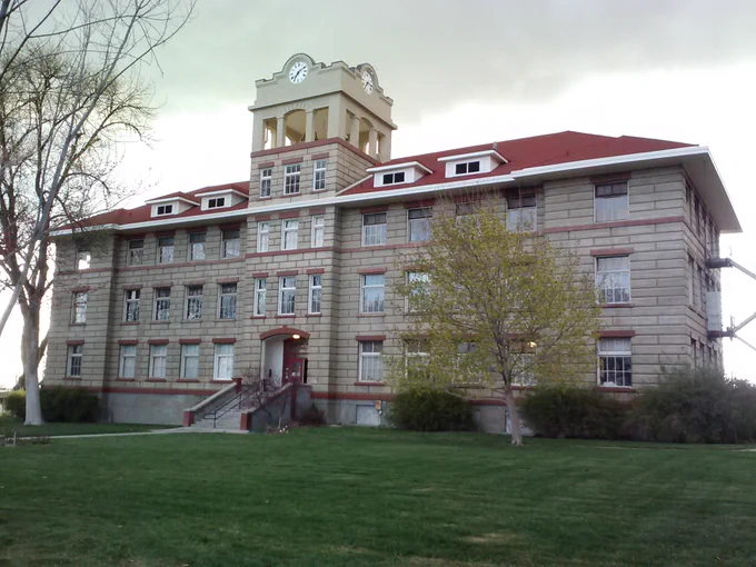  A three-story white stone building with a red roof.  In the center of the building is a taller clock and bell tower.  Steps lead to the building’s entrance, within the center section. 