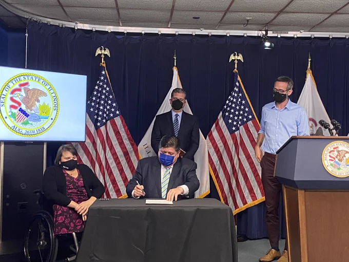 Governor Pritzker is sitting at a table signing the executive order. A white woman in a wheelchair, white man in a dark suit, and a white m an in a blue shirt all watch. Behind them are American and Illinois flags.