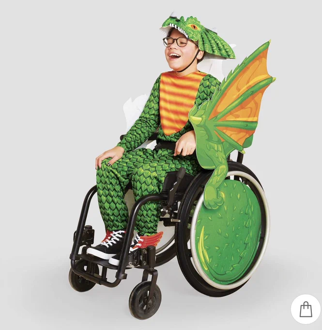 Photo: "Kids' Adaptive Dragon Halloween Costume Wheelchair Cover with Headpiece from Hide & EEk Boutique available at Target.  Photo Source Target, Young boy
smiling as he models dragon costume, green scales with green & yellow wings, yellow and orange striped chest and green headpiece/head of roaring dragon.  Wheel cover
is decorated with green scales.  He is wearing black and white high top sneakers with mini, red dragon wings.