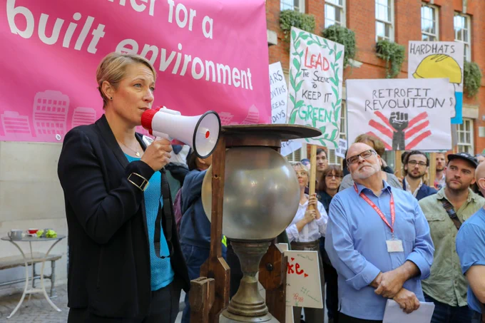 Picture is of Julie Hirigoyen, a white woman with short blonde hair wearing a blue top and black jacket. She's speaking into a megaphone in front of a pink banner at the Climate Strike. To the left of the image is a glimpse of the crowd she is talking to, who are holding protests signs. 