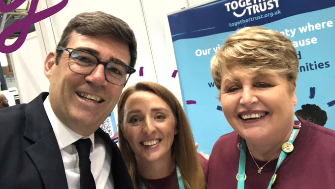 Two women, members of Together Trust staff, wearing purple Tshirts standing next to a man, the Mayor of Manchester, dressed smartly. Behind them there are colorful recruitment banners. 