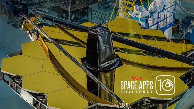 Three technicians examine the primary mirror of the James Webb Space Telescope, which is laid on the ground. A "NASA Space Apps Challenge" 10th year special logo is on the bottom right of the image.