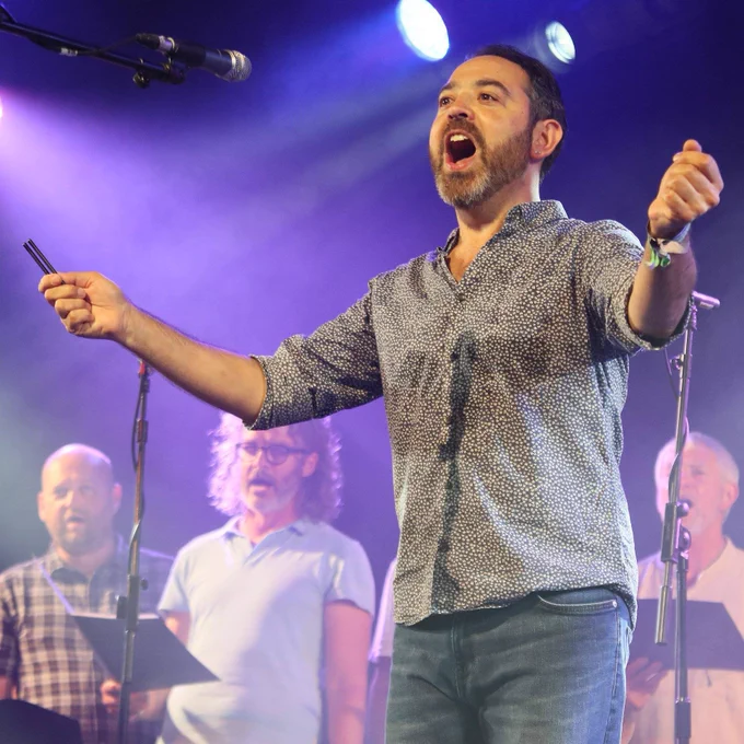 Paul Sartin singing into a microphone on stage with other singers in the background.