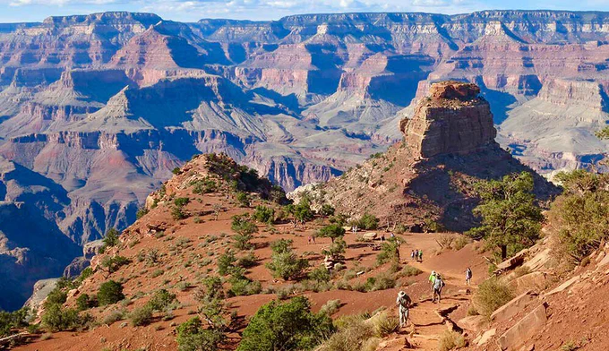 From an open terrace, hikers are ascending a hill within a colorful canyon landscape of towering peaks and cliffs. NPS/M. Quinn