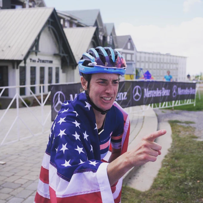 Mountain bike racer Lea Davison smiles and points as if teasingly scolding someone, wrapped in an American flag, after the finish of a race.