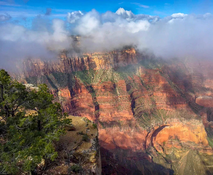 A blanket of fluffy clouds across the horizon above layer upon layer of colorful rocks creating an imposing cliff several thousand feet high. NPS Photo/M Quinn