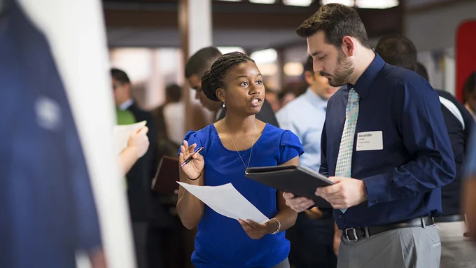 A female recruiter dressed in blue looks over a resume while speaking with a male student dressed in navy.