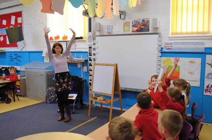 Isabelle King stands in a classroom, her hands held up high. In front of her, a row of school children enthusiastically return the gesture.