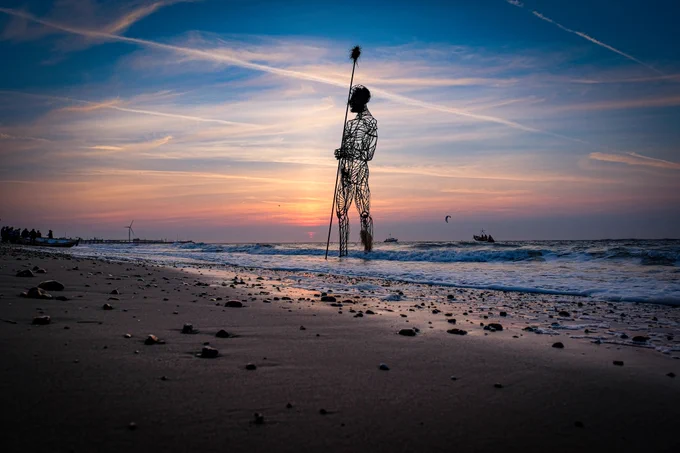 A dramatic beach photo shows a vast sculpture of a man standing against a beautiful sunset.