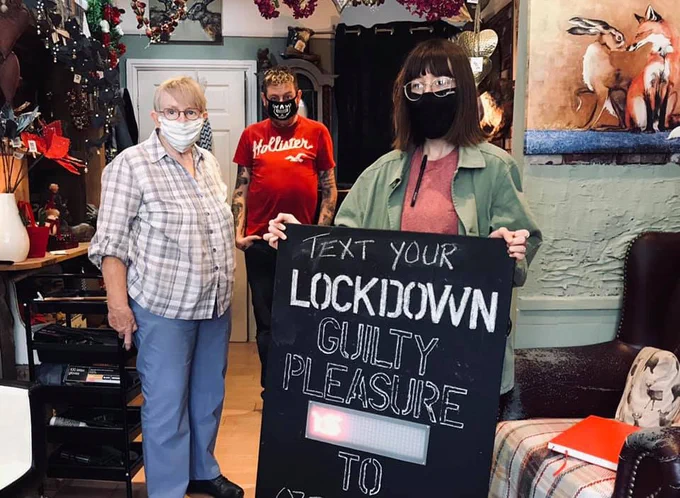 Emily Godden stands in a busy environment, holding a chalkboard that reads 'Text your lockdown guilty pleasure to', with the bottom of the sign cut off. 