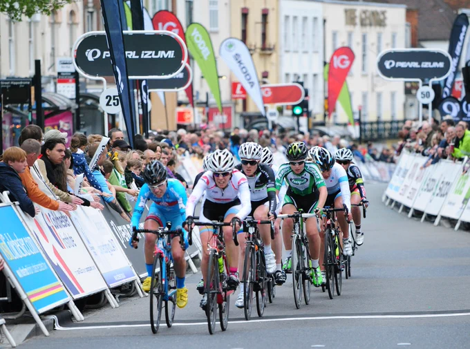 A photo of the Tour Series in Colchester in 2013. The photo shows the riders cycling towards the camera, up the High Street. Either side of the riders are people lining the streets cheering them on and watch them go past.