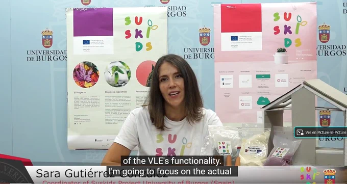 Sara G. from UBU sit behind a desk, with a SUSKIDS tshirt, explaining. Behind her a wall with SUSKIDS posters and on the right of the image a house made in cardboard with bags of different  powder that are building materials