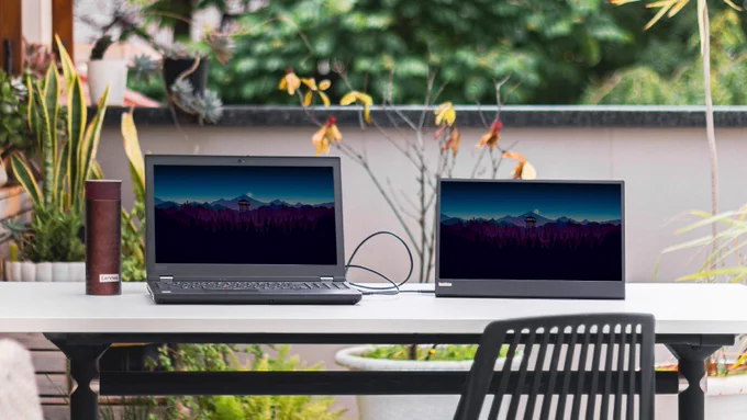 A large brown Lenovo coffee cup and 2 Lenovo laptop screens on a grey table outside.
