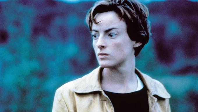 A woman looks out beyond the distance. She has short hair, and is wearing a black shirt underneath a yellow jacket.