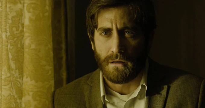 A bearded man in a white shirt and blazer stands by a window. He looks concerned. Beside him are yellow curtains.