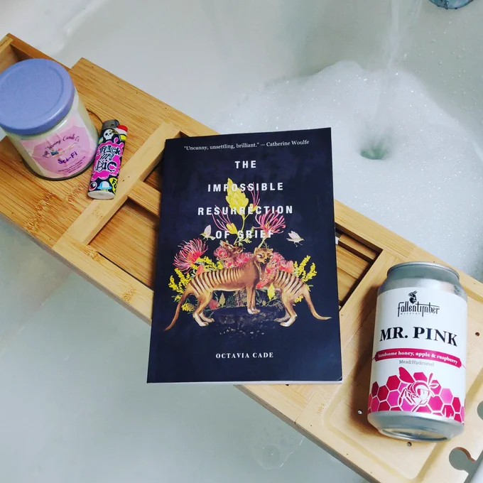 A board across a tub that is filling with water and bubbles holds a candle, lighter, paperback book, and can of mead.