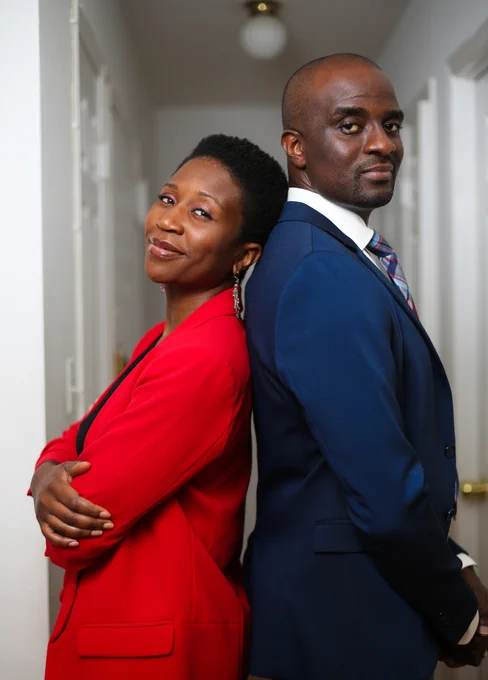 Black woman in red suit and Black man in blue suit standing back to back