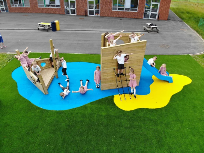 several children in red school uniform pose and look up to the drone camera on one of our imagination station products which is seated on blue and yellow wetpour splashes and green artificial grass surfacing 