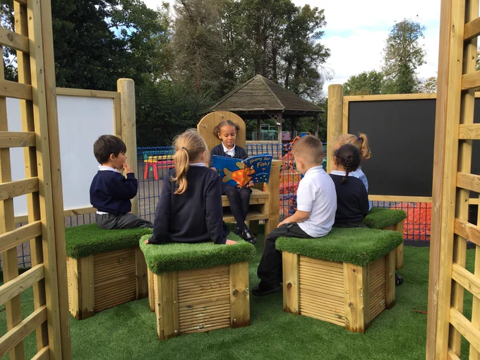 a child sits on our storytelling chair, reading a story to five of her classmates as they sit on artificial grass topped seats