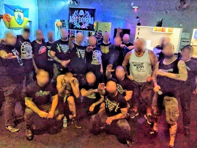 Foto de vários homens brancos com o rosto censurado fazendo a saudação nazista (braço esticado para frente). Quase todos usam camisetas com símbolos neonazistas. Ao fundo é possível ver uma bandeira do Batalhão Azov