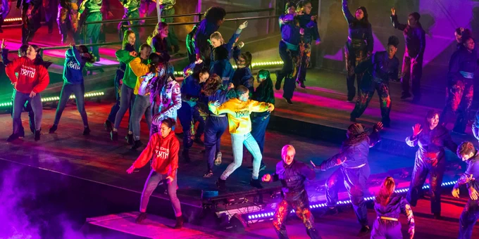 A crowd of young people dancing on a long stage, wearing jeans and different coloured tops, facing different directions and in dynamic poses, with colourful lights and smoke effects around them.