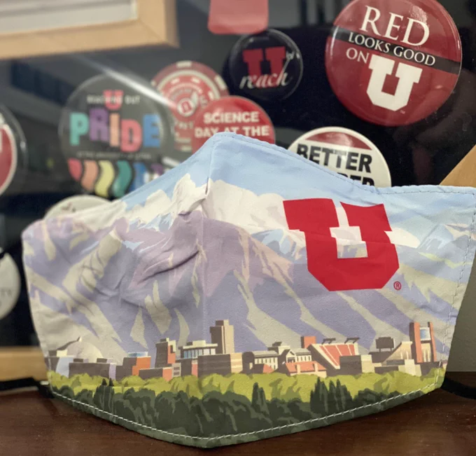 Photo of a facemask, which has an illustration of the University of Utah campus on it, set against a button board of past U pins.