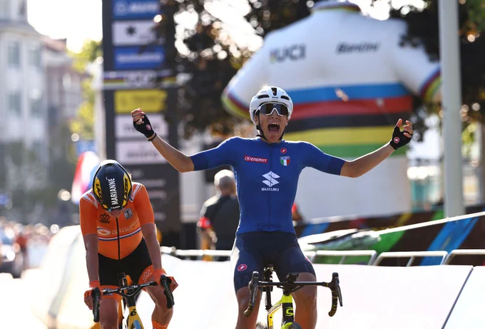A photo of cyclist Elisa Balsamo winning the World Championship in Leuven. She's raising her hands in the air, cycling and shouting in joy. Behind her is another rider cycling towards the end of the race.