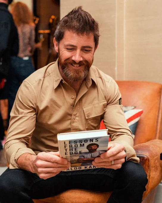 Photo of Aldo Kane smiling and holding a copy of his book: Lessons From The Edge.