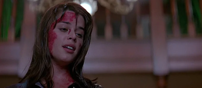 Neve with blood covering the left side of her forehead and face. The camera angle is low and she is looking down at someone with a relieved look and a slight smile.