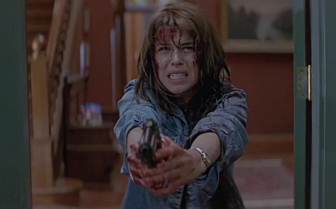 She is stood at the front door and the shots point of view is of someone on the doorstep of the house. She is covered in blood holding a gun in front of her with both her hands. She seems to be crying with a pained expression on her face.