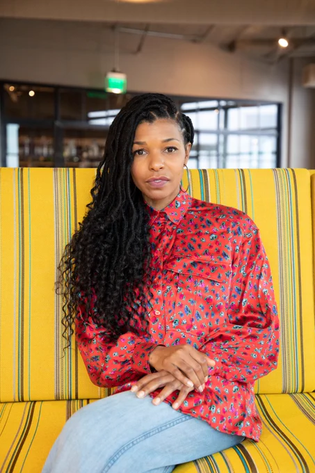 Photo of Kimberly McGlonn, a Black woman in a red shirt and blue jeans, sits on a yellow striped couch.