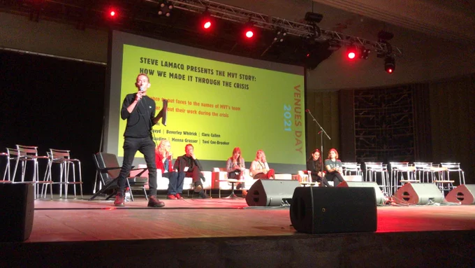 Steve Lamacq dressing in black stands on a stage holding a microphone in front of a panel of people.