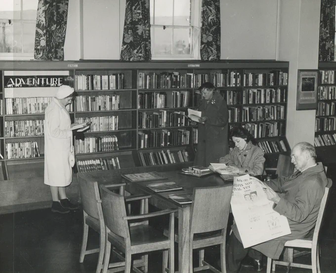 An old photo of the interior of Cheriton Library. Customers read newspapers at a table while other;s browse the adventure section