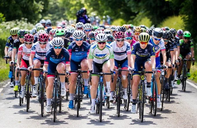 A photo taken of a women's cycling race, taken from the front as the riders head towards the camera, down a tree-lined road with hundreds of riders blurring into the distance.