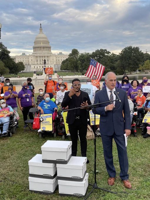 Senator Bob Casey stands on the grass in front of the US Capitol, speaking into a mic with 5 large white boxes to his left. Behind him are dozens of advocates holding signs.