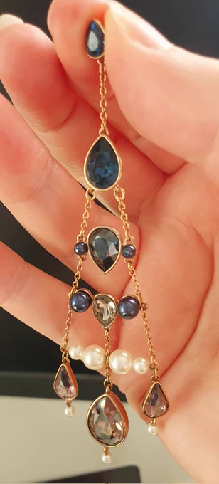 A very long copper coloured earring with light and dark grey crystals in a triangle shape, near the bottom is a row of white pearls.