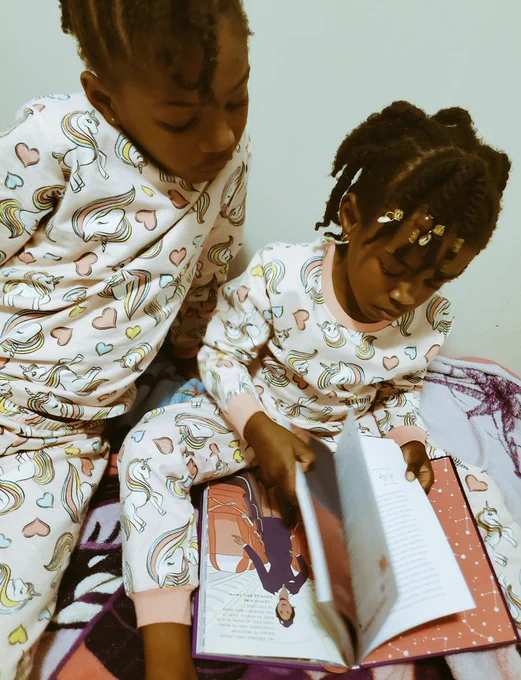 Two little Black girls in matching pink unicorn pj's, reading a book