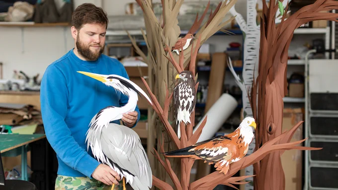 A person holds a model of a bird while standing in front of model trees and other small birds