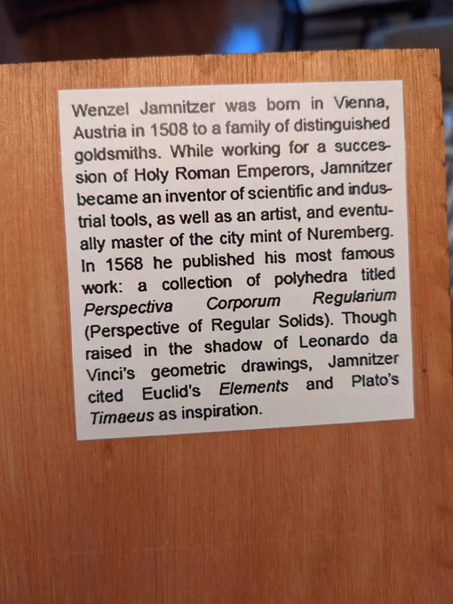 The back of the wood, which reads, "Wenzel Jamnitzer was born in Vienna, Austria in 1508 to a family of distinguished goldsmiths. While working for a succession of Holy Roman Emperors, Jamnitzer became an inventor of scientific and indus trial tools, as well as an artist, and eventually master of the city mint of Nuremberg. In 1568 he published his most famous work: a collection of polyhedra titled Perspectiva Corporum Regularium (Perspective of Regular Solids). Though raised in the shadow of Leonardo da Vinci's geometric drawings, Jamnitzer cited Euclid's Elements and Plato's Timaeus as inspiration."