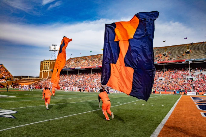 two cheerleaders running with huge Block I flags across the field