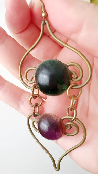 A long gold wire earring in 2 parts, in the top curled shape is a clear green bead, in the bottom curled shape is a violet bead.