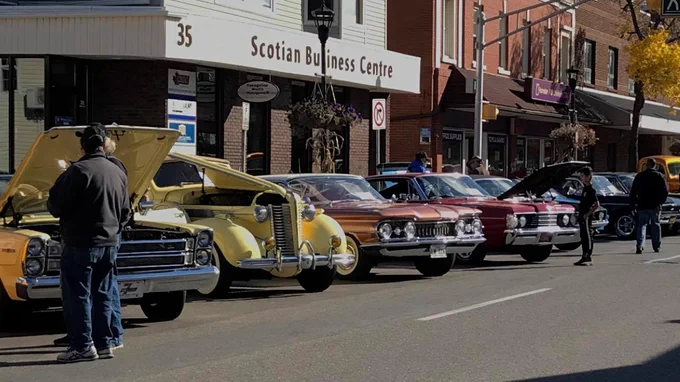 Six classics cars lined up along the street,  two of which have the hoods popped to show their engines. Four people are looking at the cars. 