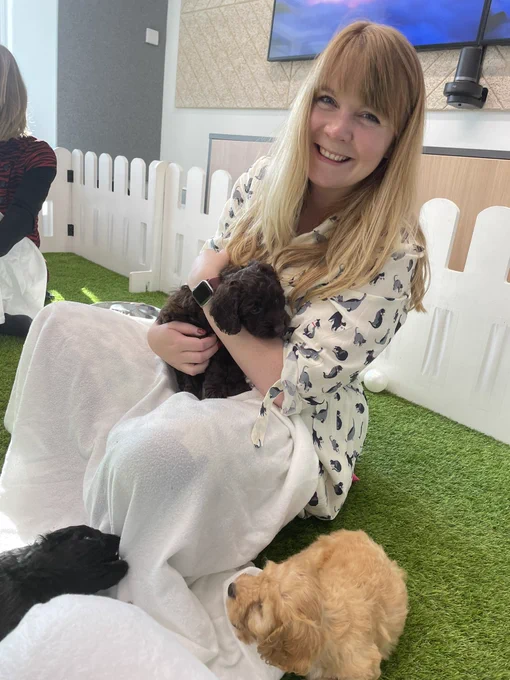 Caroline smiling while holding a chocolate brown cockapoo puppy while a caramel coloured one sits next to her and a black one nibbles a white blanket on her lap. Sat on green astroturf with a white picket fence in the background.