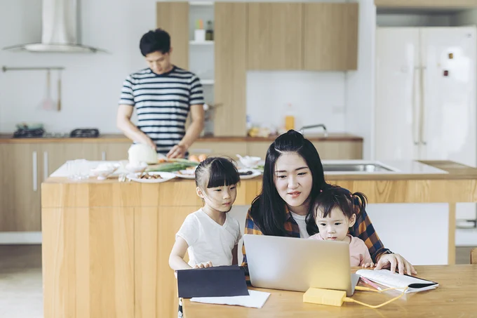 mom and daughters working on a laptop at the kitchen table while dad is preparing food in the kitchen behind them.