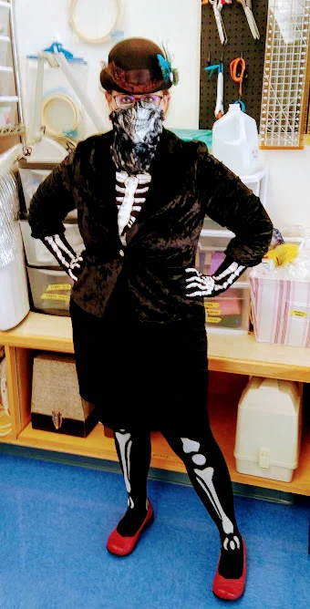 Woman in a skeleton costume with a fancy blazer and hat and red shoes, standing in front of a display of sewing equipment.