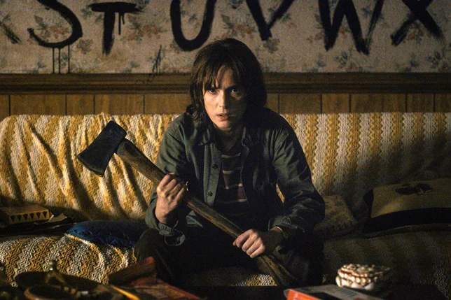 Winona in Stranger Things, she sits cross legged on her couch holding an axe.
