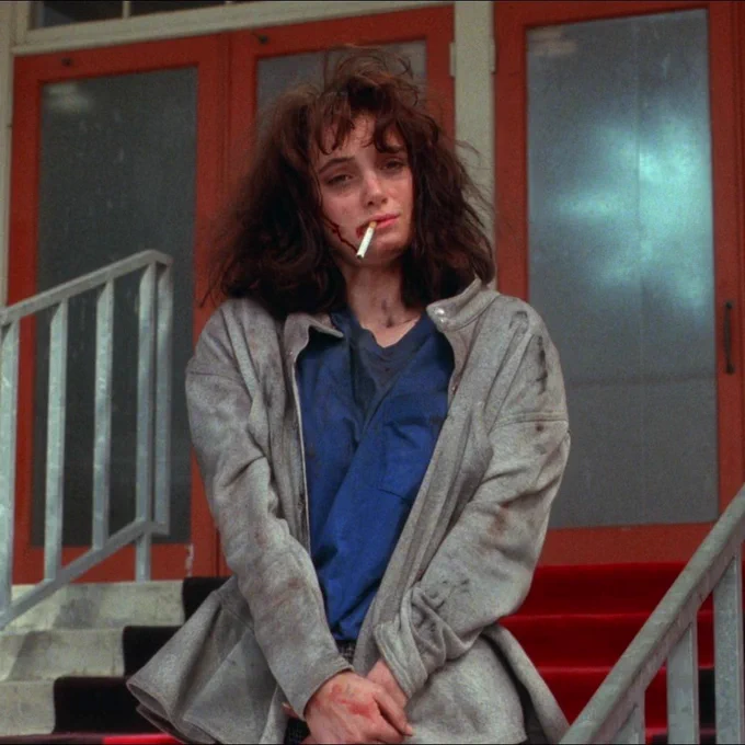 From Heathers. Her clothes and face are dirty with her hair messy. She has a cigarette hanging from her mouth and looks slightly dazed.